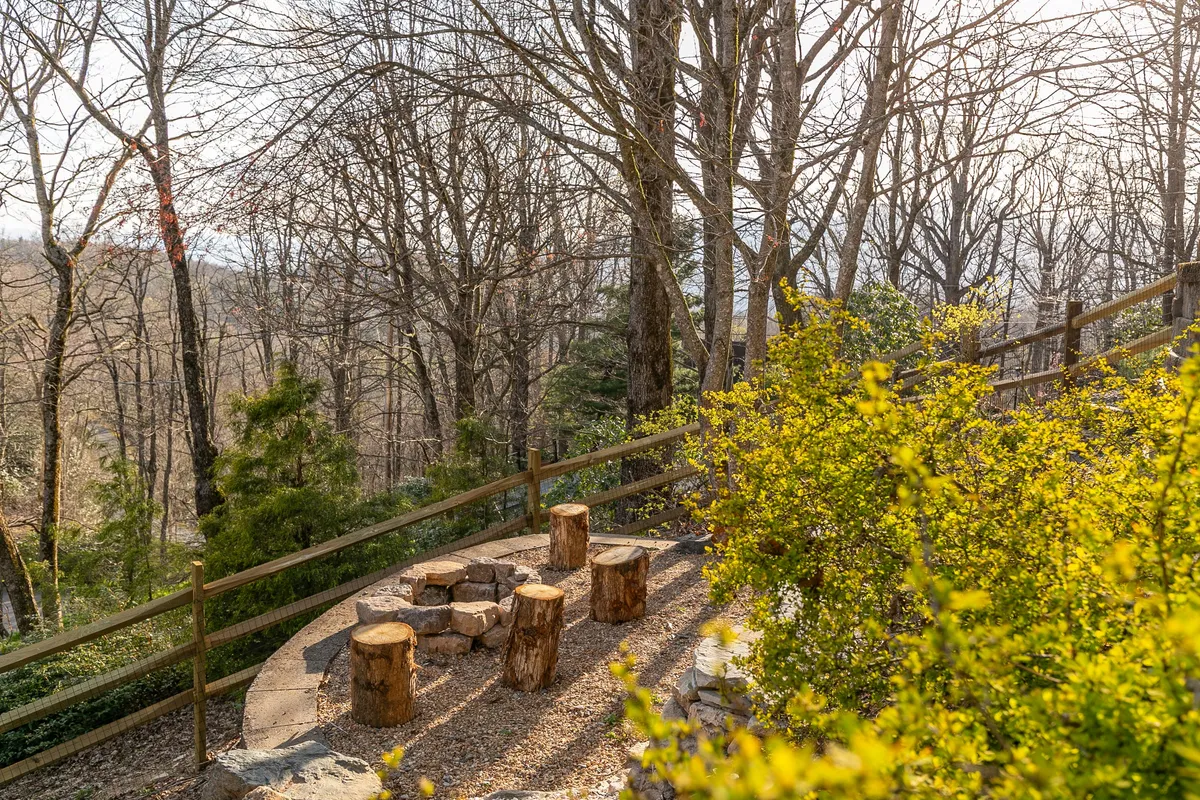 A serene fire pit area with rustic seating logs arranged around a gravel circle, surrounded by tall trees and natural greenery. The scene suggests evenings spent stargazing, roasting s'mores, and sharing stories by the fire.