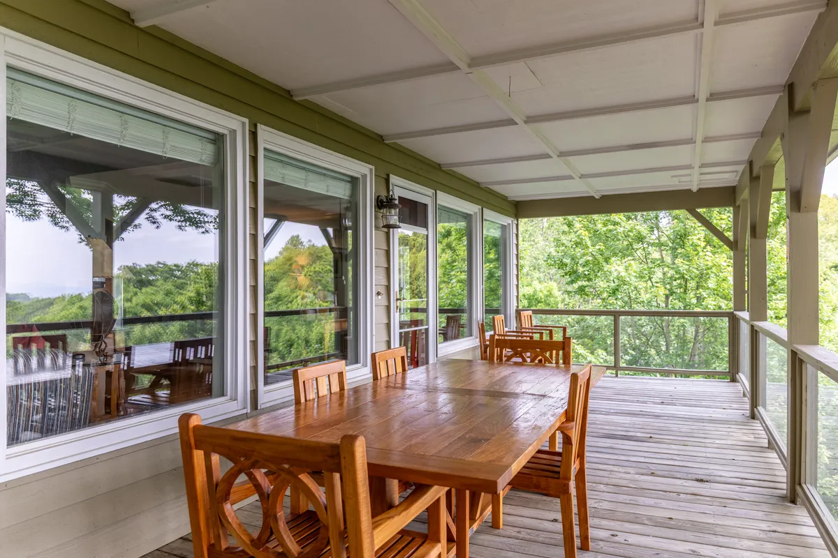 A deck with a wooden dining table surrounded by natural greenery, with large windows reflecting the ambient light. The space is set for dining al fresco or morning coffee, with mountain and sunset views completing the tranquil mood.