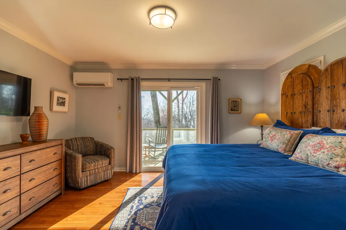 A spacious bedroom with a large blue bed, wooden dresser, comfortable chair, and a sliding glass door that leads to a private outdoor space. The room is filled with natural light, suggesting easy access to fresh air and peaceful views.