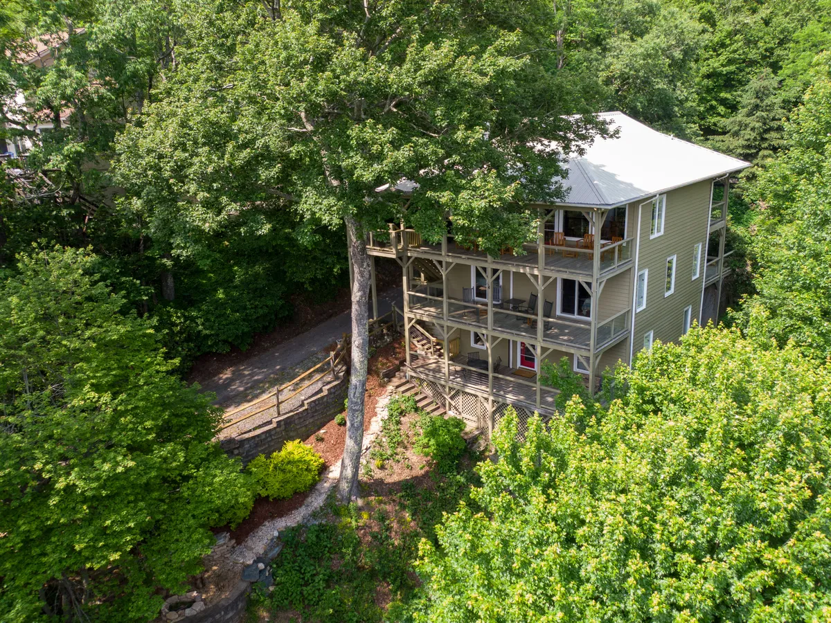 This image showcases the home’s multi-level deck nestled under a large tree, with inviting pathways leading toward the house. On the deck, people are seen relaxing and gathering for meals, all set against a backdrop of sunset views, lush greenery, and hints of the fire pit area below.