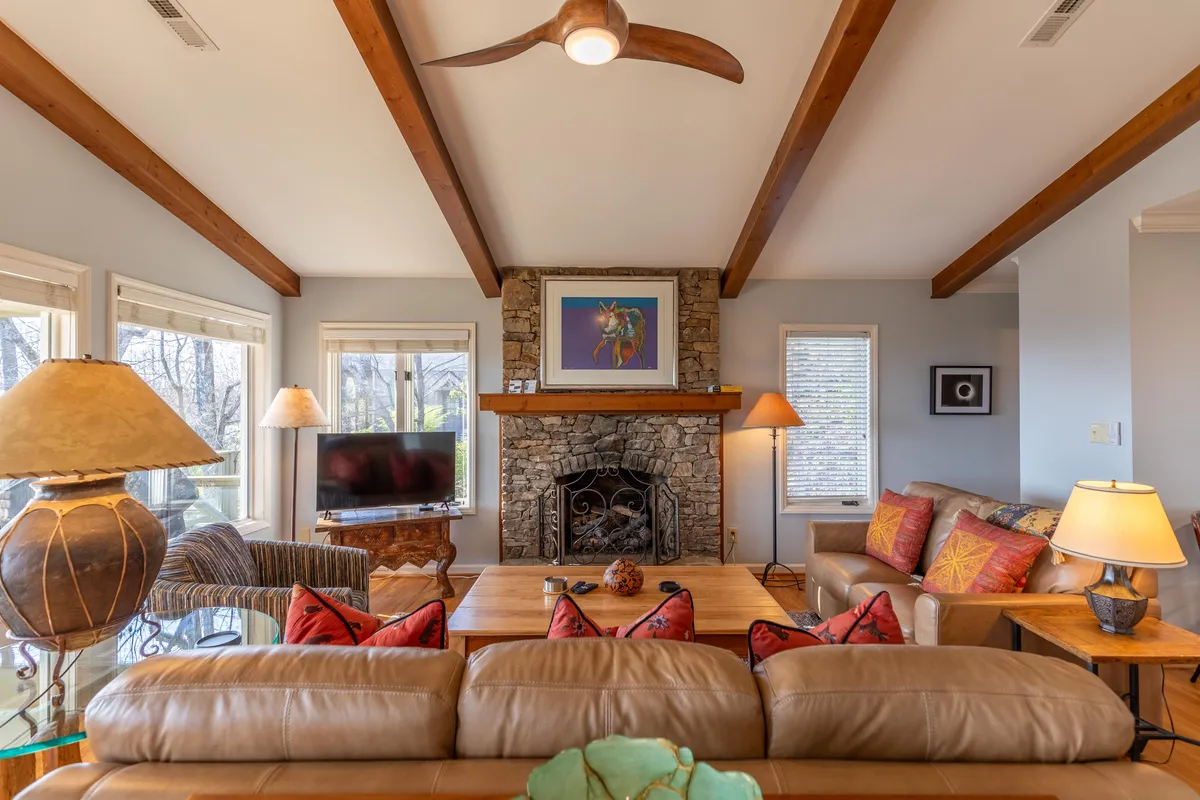 A warm, open-concept living room centered around a striking stone fireplace. The room features exposed wooden beams, plush seating, colorful artwork, and large windows that let in abundant natural light. Decorative lamps and cozy textures create an inviting environment perfect for gatherings.