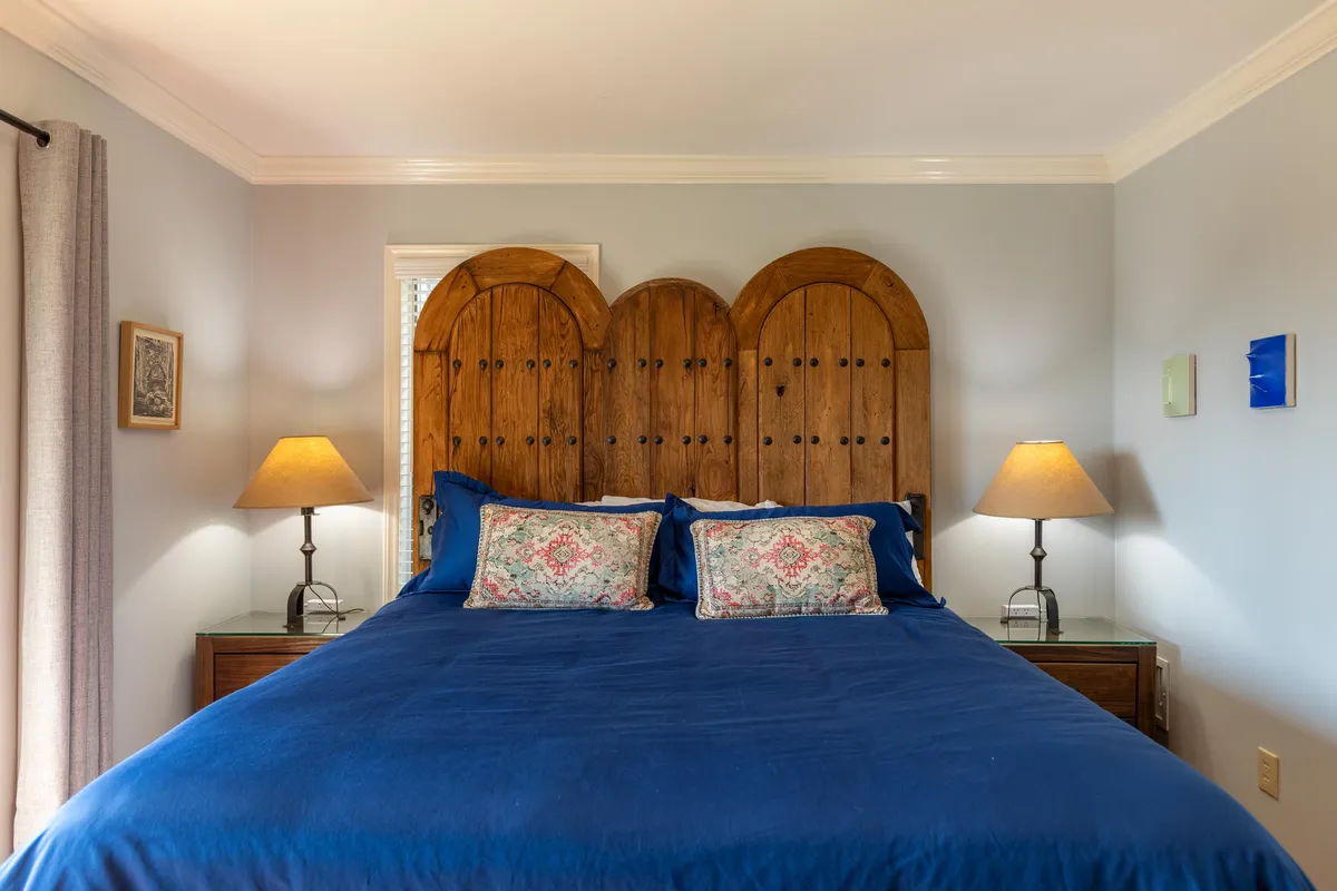 A serene and cozy bedroom featuring a wooden headboard, blue bedspread, decorative pillows, and nightstands with lamps. Natural light filters in, creating an inviting space perfect for relaxing, reading, or enjoying a quiet moment.