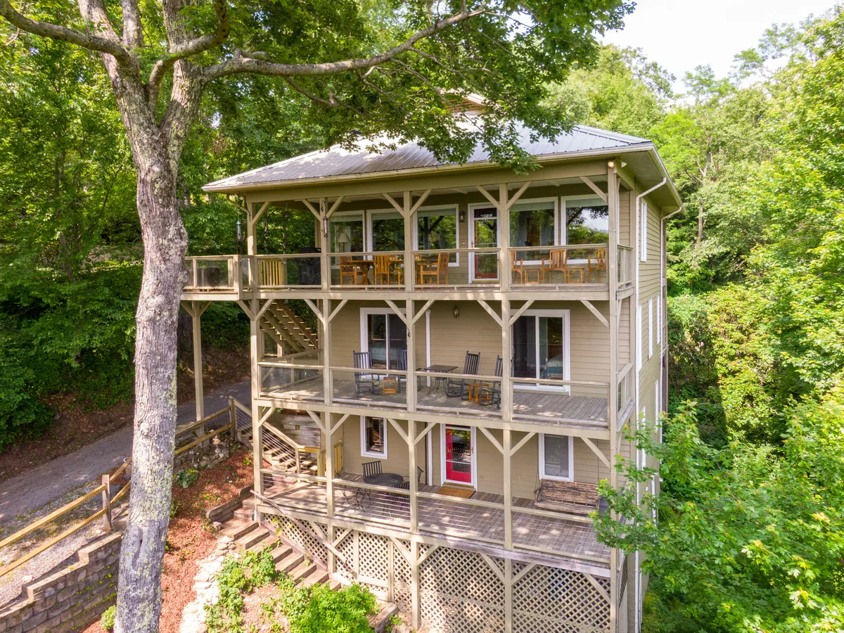 The image showcases a beautiful multi-story house nestled among lush greenery in the Misty Mountain community of Blowing Rock. The home features inviting balconies with comfortable seating, perfect for soaking in the mountain views. A large tree frames the scene, adding to the serene and private atmosphere, while the expansive outdoor spaces hint at opportunities for relaxing, dining, and family gatherings.