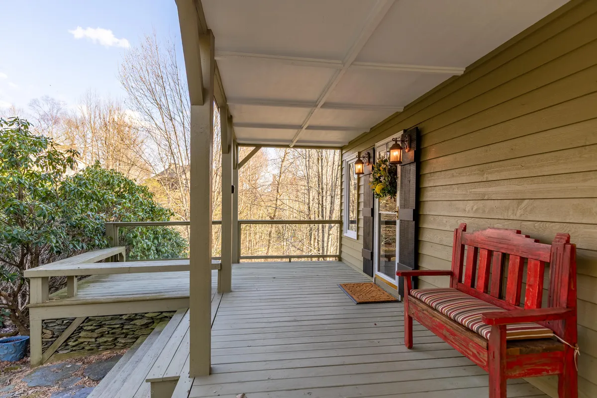 This image features a charming exterior view with a red bench on the porch, a welcoming doorway adorned with decorative lights, and lush greenery framing the entryway. The setting invites outdoor relaxation and hints at cozy gatherings on the porch, connecting the home to its scenic natural surroundings.