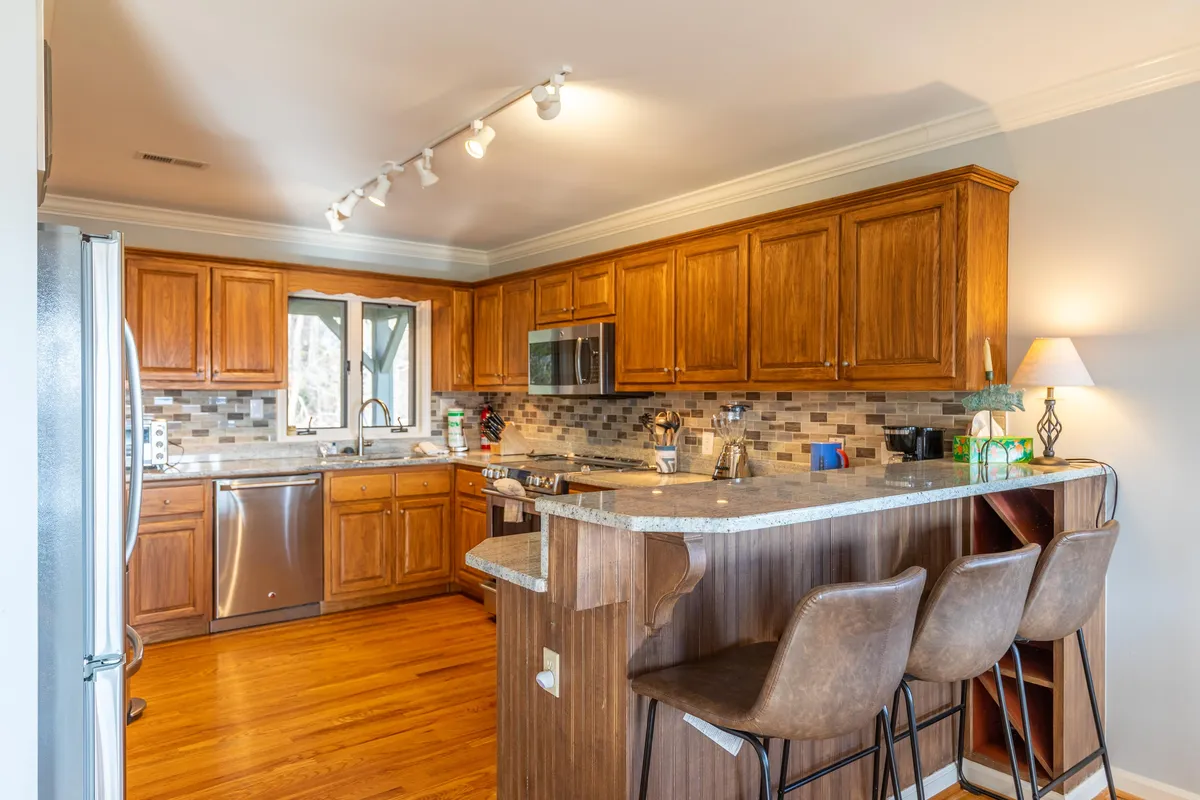 A well-appointed gourmet kitchen featuring rich wooden cabinetry, gleaming granite countertops, stainless steel appliances, and bar stools for casual dining. The space is designed for both culinary creativity and social connection, with ample room for preparing feasts or enjoying a morning coffee.