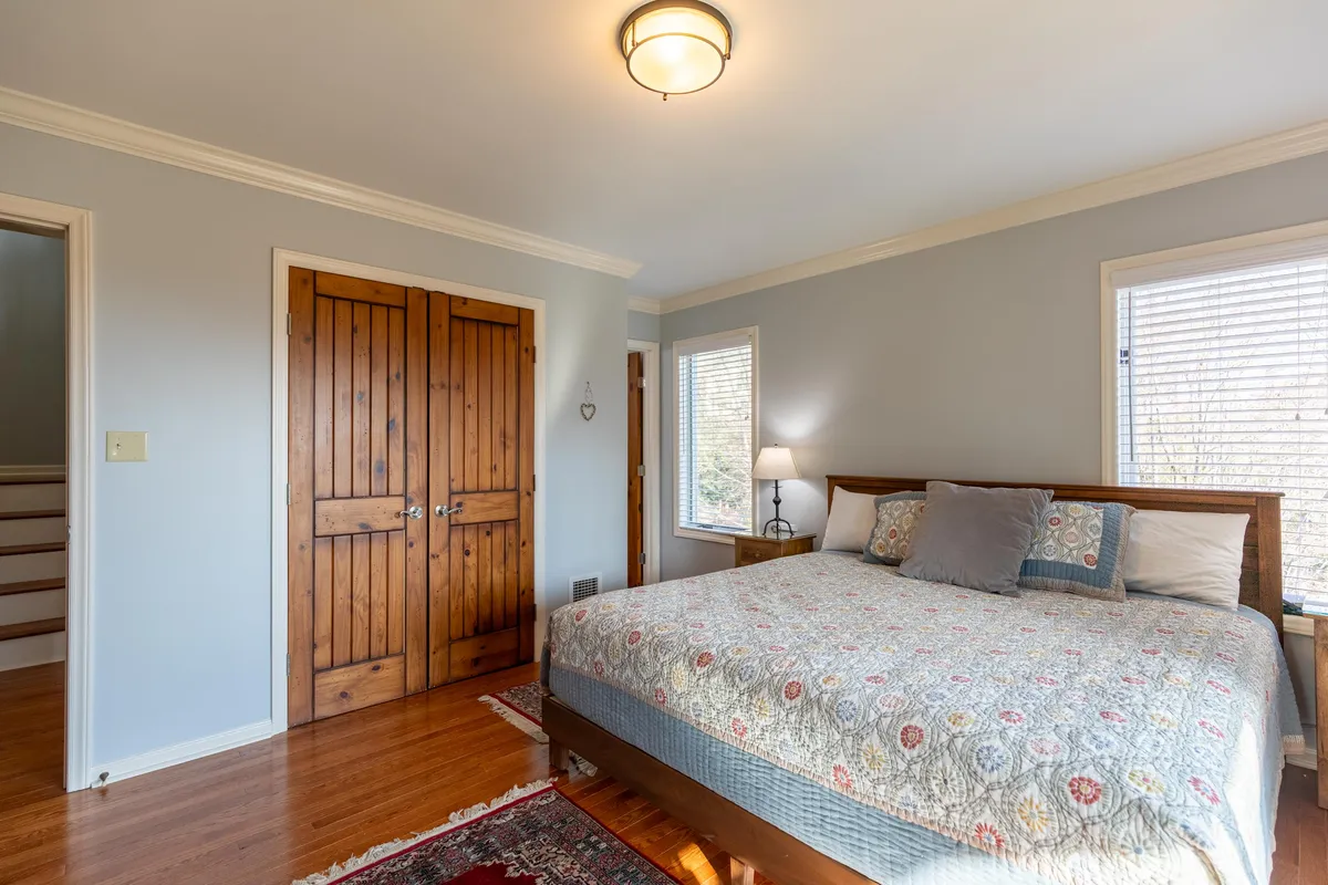 A tranquil bedroom featuring a bed with patterned bedding, wooden doors, and a window letting in abundant natural light. The decor is simple yet inviting, and the soft textures create a relaxing environment ideal for restful nights or afternoon reading.
