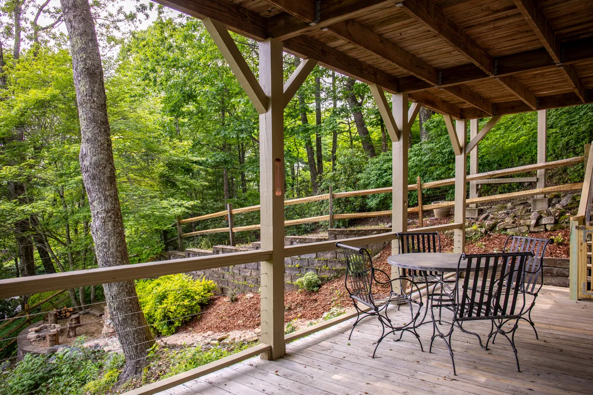 An inviting exterior shot of a wooden deck featuring a metal table and chairs, surrounded by lush greenery and a stone wall. The deck is designed for outdoor dining, relaxation, and enjoying the natural landscape, suggesting both privacy and opportunities for shared moments with a mountain backdrop.