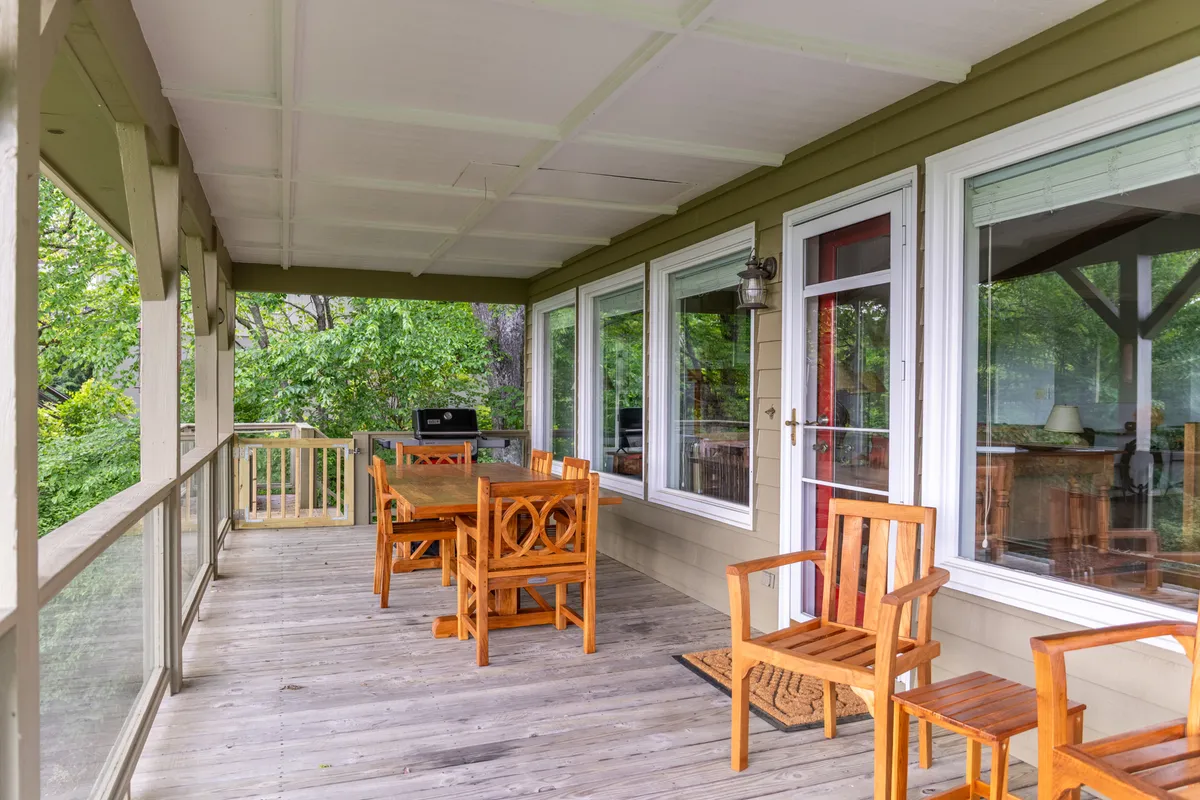 A spacious deck furnished with wooden tables and chairs, large windows facing out to greenery, and hints of mountains in the distance. The setting is perfect for outdoor meals, relaxation, and socializing while enjoying panoramic sunset views.